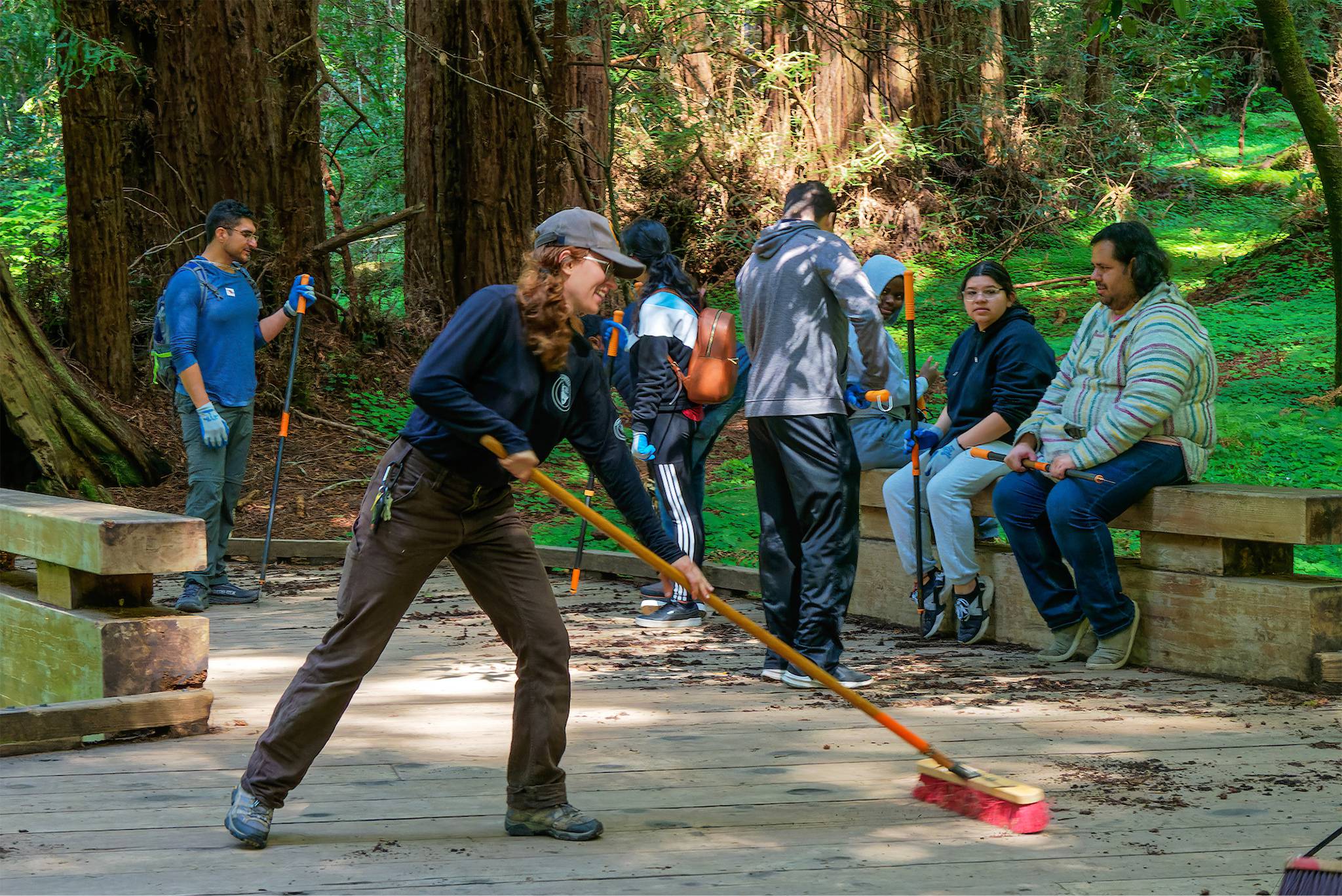 A female volunteer smiles as she sweeps a deck near redwood trees as other volunteers pause to sit on nearby benches