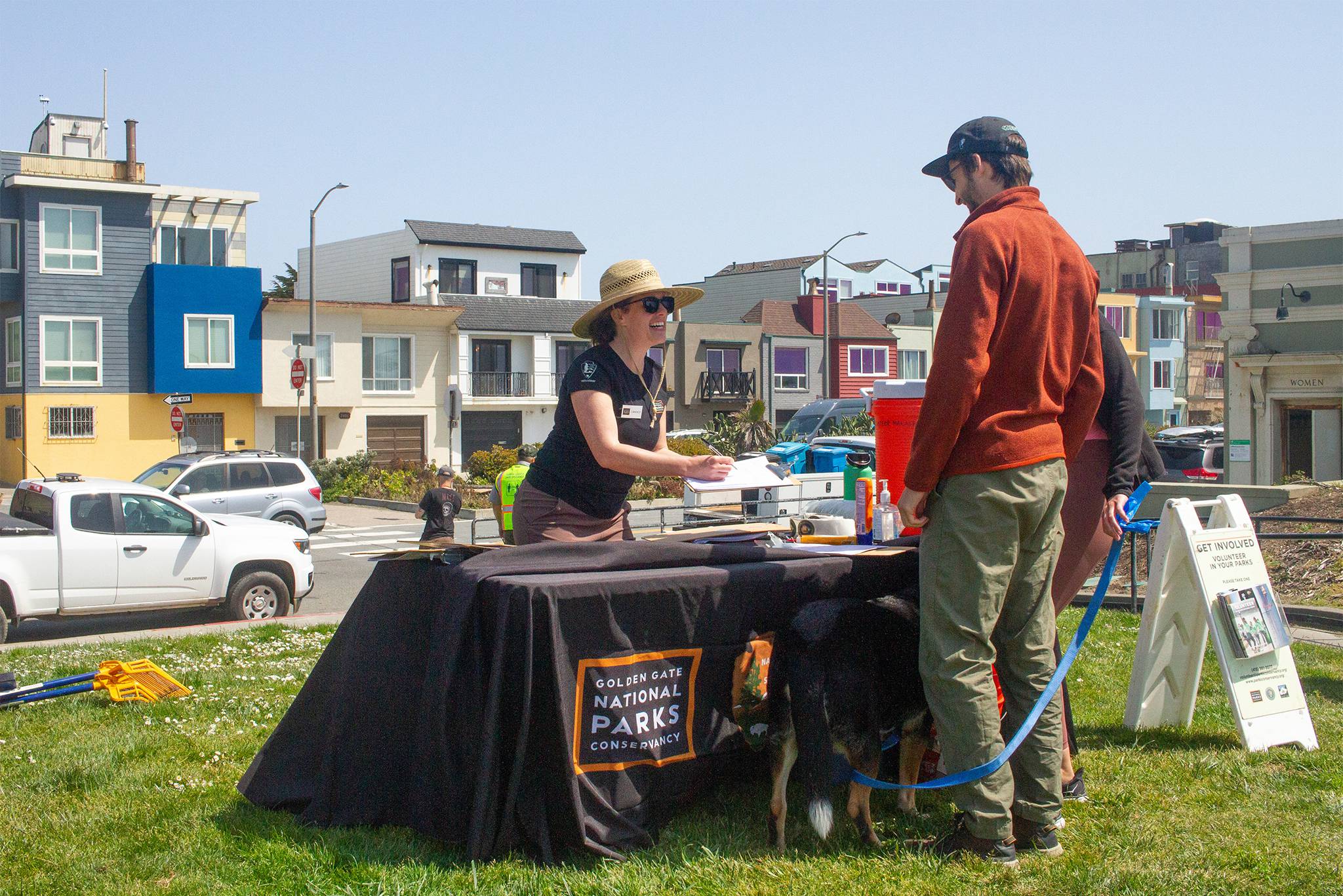 Standing behind a GGNPC table on the grass at Crissy Field, a smiling female volunteer in straw hat and sunglasses holds a clipboard and speaks to a couple and their dog