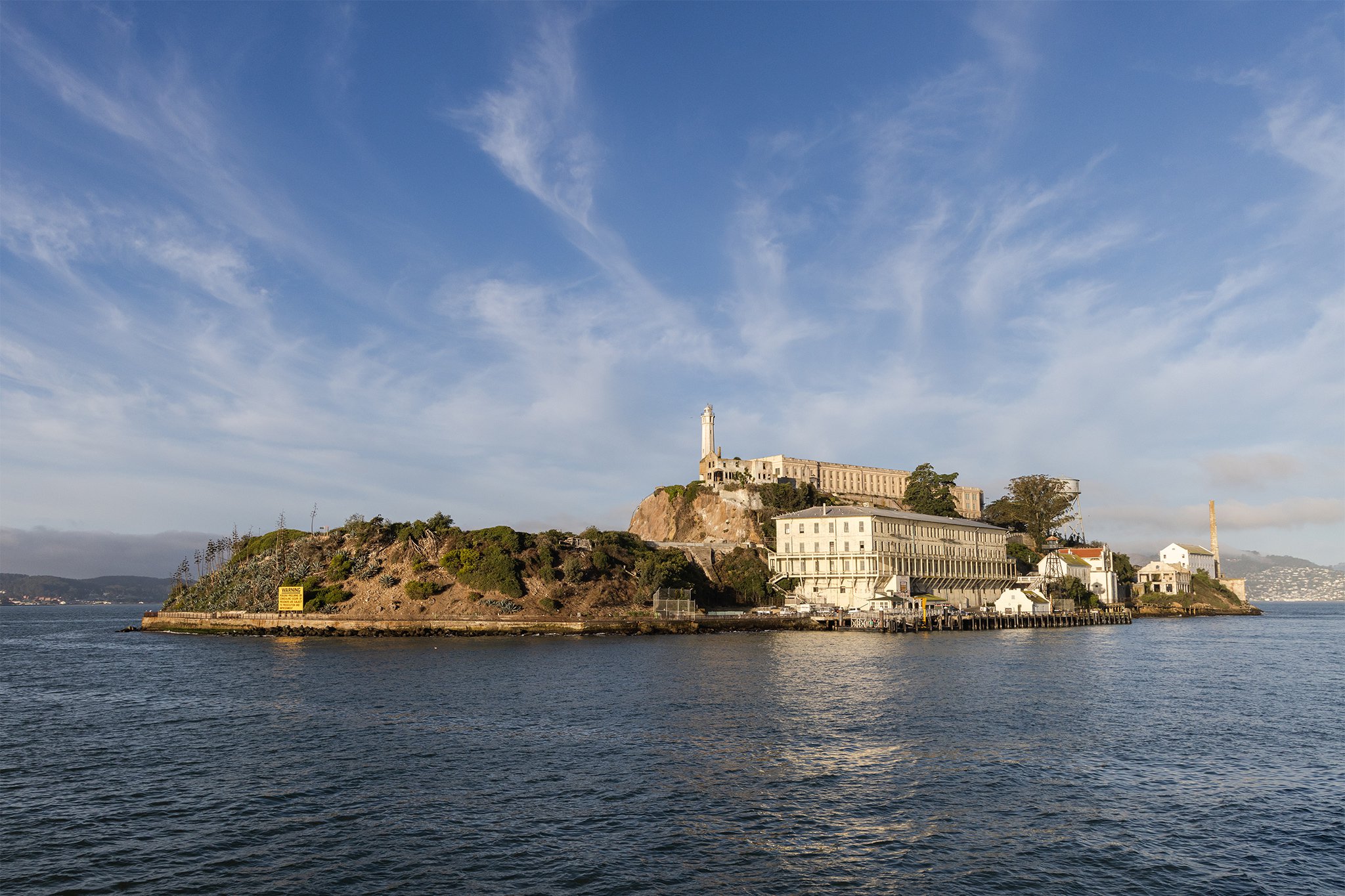 From aboard an Alcatraz Cruises boat, a sunny view of Alcatraz Island, the waters of SF Bay and blue sky with wispy clouds.